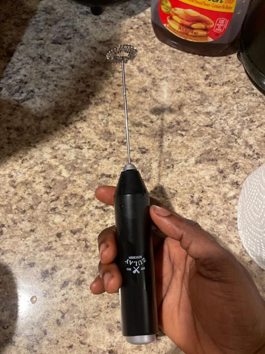 A hand holding a black handheld milk frother with a whisk attachment on a kitchen countertop.