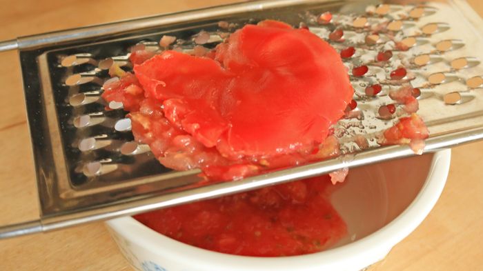 Tomato skin rests on a grater, with the tomato pulp collected in a bowl beneath it.