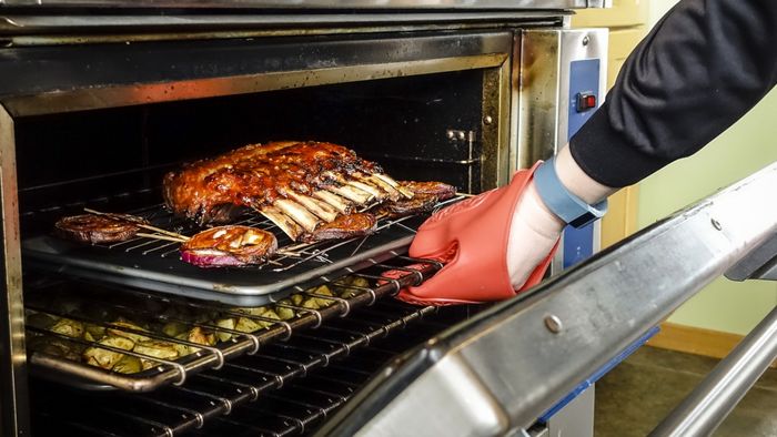 A hand, clad in an oven mitt, reaches into a blazing oven to retrieve a rack of ribs.