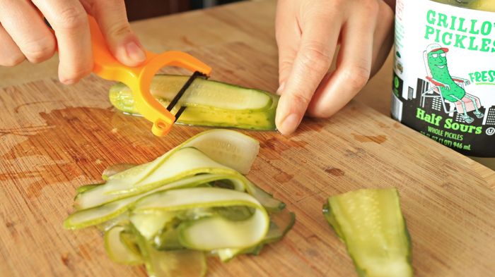 Using a vegetable peeler, hands gently shave off thin slices from a pickle.