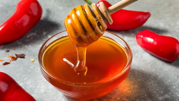 A honey dipper drizzling golden honey into a bowl, with vibrant red peppers in the background.