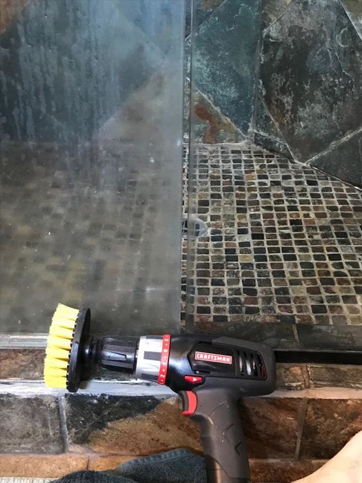 A drill-mounted scrub brush is placed next to a shower pane, with one side still foggy and the other sparkling clean.