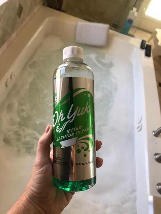 A hand holding a bottle of Oh Yuk Jetted Bathtub Cleaner in front of a bathtub full of water
