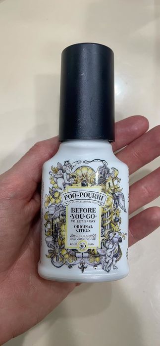 A hand holds a bottle of Poo-Pourri's original citrus-scented toilet spray. The bottle features a decorative label with floral and bird illustrations.