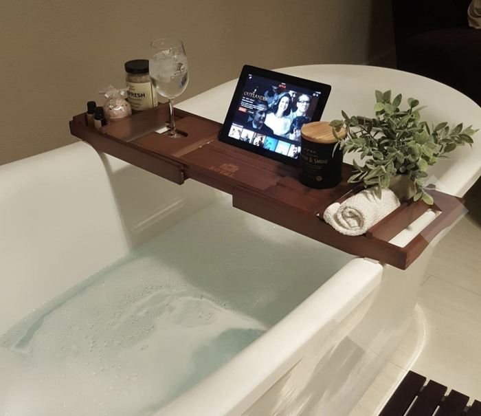 A dark wood bamboo caddy placed over a bathtub, holding an iPad, a candle, and other accessories, creating a serene and organized bath setting.