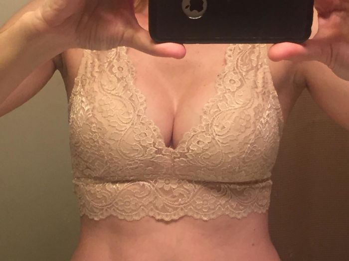 A person takes a mirror selfie while wearing a lace bralette, with their face not visible in the shot.