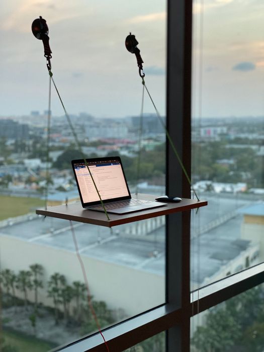 A wooden desk suspended by ropes and suction cups, mounted to a window with a laptop placed on it.
