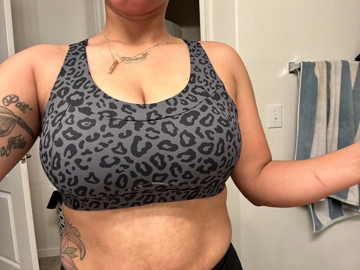 A reviewer taking a selfie from the upper chest to waist, wearing a leopard-print sports bra. Tattoos are visible on her arm and a floral one on her lower abdomen.