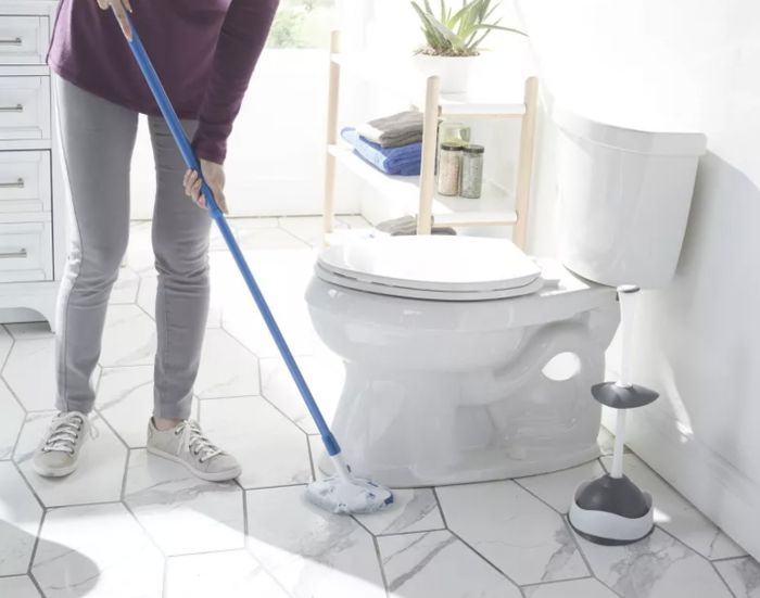 A person using an extendable pole scrubber to clean the area beneath a commode