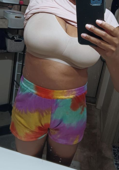 A person in a beige bra and tie-dye shorts taking a mirror selfie, with their head not visible in the photo