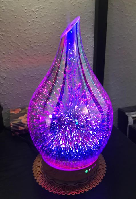 A bulb-shaped glass oil diffuser illuminated with purple and blue lighting effects reminiscent of fireworks.