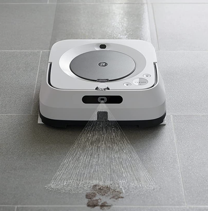The iRobot vacuum spraying a cleaner on a floor stained with muddy footprints.