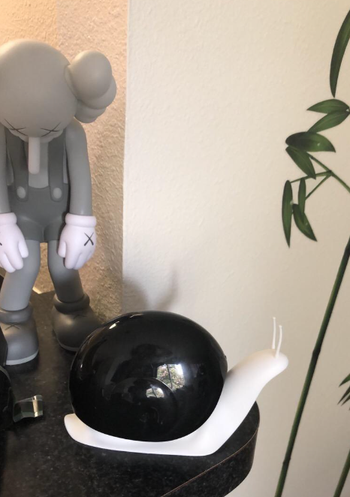 A minimalist snail-shaped soap dispenser placed on a display table surrounded by a figurine and a plant.