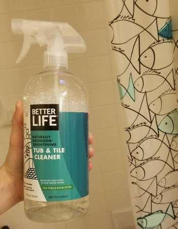 A hand holding a bottle of Better Life bathroom cleaner with a shower curtain backdrop