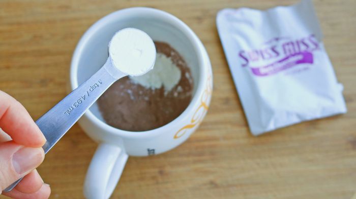 A hand holds a teaspoon of cornstarch above a mug filled with hot cocoa mix.
