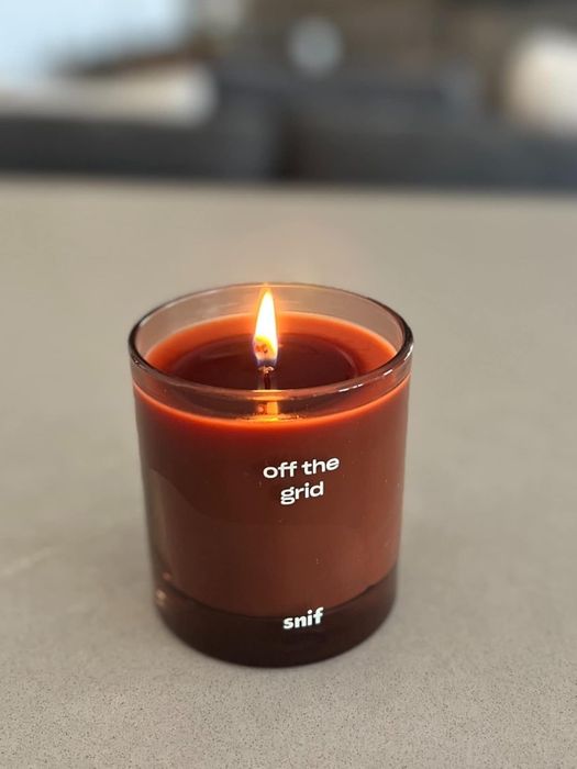 A lit scented candle in a glass jar labeled 'Off the Grid' by Snif, sitting on a smooth surface.