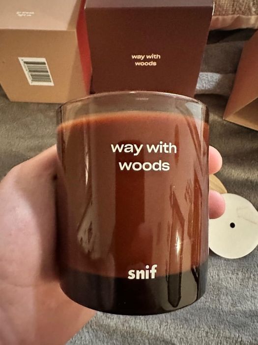 A hand holding a candle labeled 'Way with Woods' by Snif, with boxes in the background.