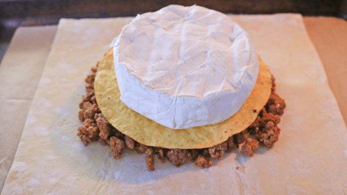 A brie wheel resting on a tortilla with seasoned ground beef layered on puff pastry.