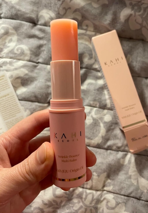 A hand holding the Kahi Wrinkle Bounce Multi Balm against a backdrop featuring its packaging.