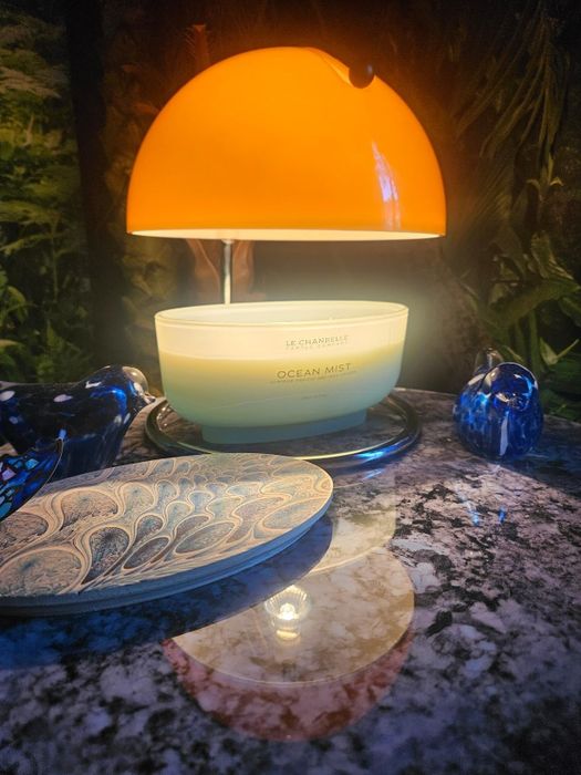 A stylish display features a glowing dome-shaped lamp, an ocean mist-scented candle, two blue ceramic birds, and an intricately patterned plate resting on a marble surface.