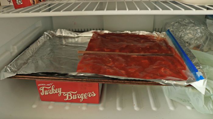 A metal tray in the freezer, with a bag of tomato sauce and a chopstick dividing it into two portions.