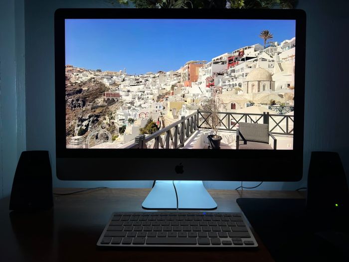 A computer screen shows a scenic hillside town with white buildings and a clear sky. The desk features a keyboard and speakers.