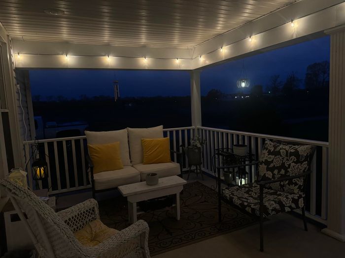 A covered porch at night, adorned with string lights, a wicker chair, a cushioned loveseat with yellow pillows, a small table, and a black armchair with floral cushions.