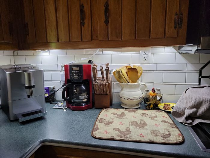 A kitchen countertop adorned with a coffee maker, a knife block, a utensil holder, a decorative rooster-themed mat, and a ceramic rooster figurine, all illuminated by under-cabinet lighting.