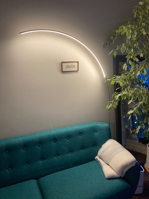 A modern living room featuring a teal tufted sofa, a cozy white blanket, a stylish curved floor lamp, and a vibrant potted plant. A striking art piece hangs on the wall behind the sofa.