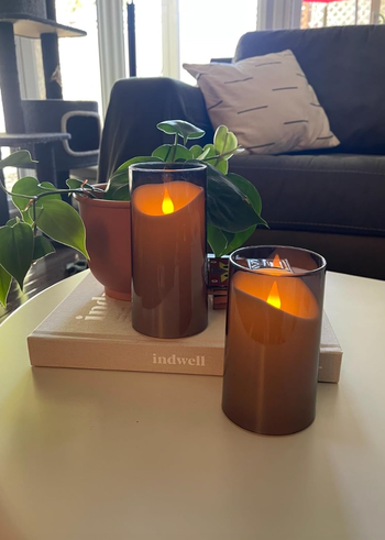 Two candles glowing in brown glass holders placed atop a book, with a plant and a sofa visible in the background.