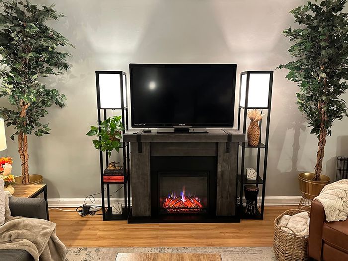 A cozy living room arrangement featuring a TV on a stand with an electric fireplace beneath it, framed by two floor lamps and lush greenery. A gray sofa sits on the left, while a basket filled with blankets rests on the right.