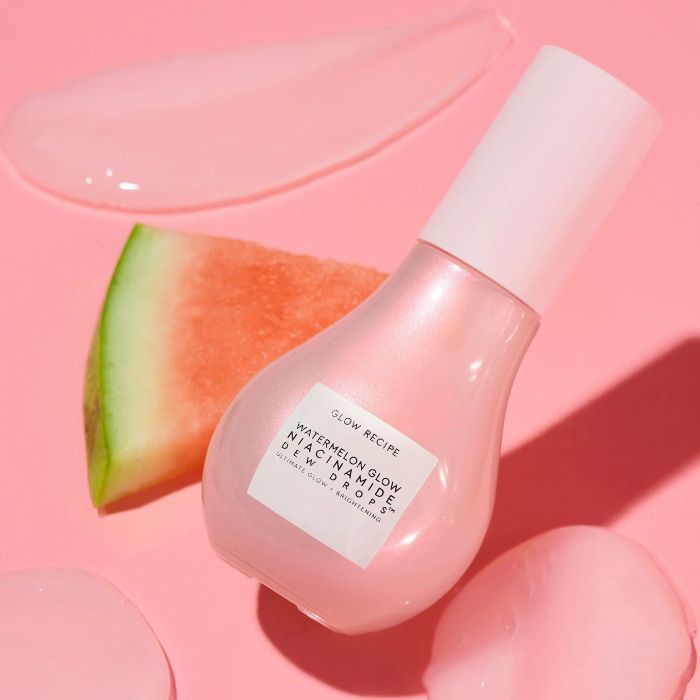 A bottle of Glow Recipe Watermelon Glow Niacinamide Dew Drops placed next to a watermelon slice on a pink background, emphasizing its dewy finish