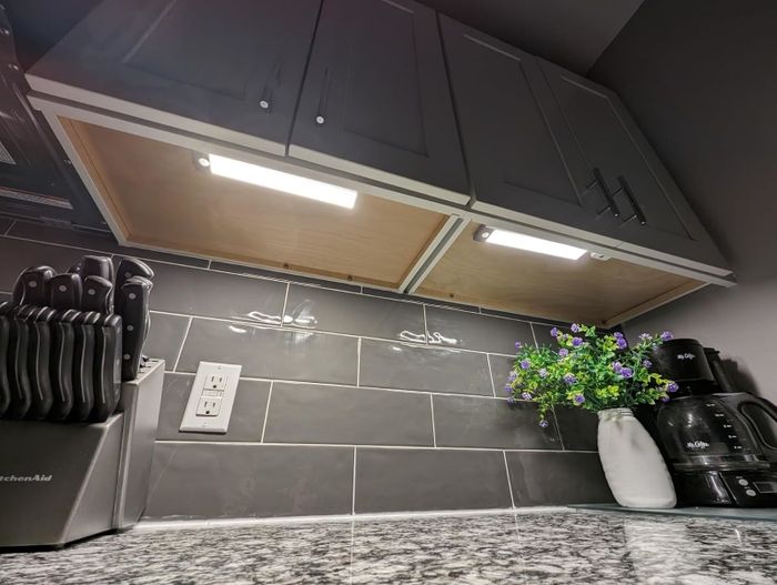 A kitchen countertop complete with a knife block, an electrical outlet, under-cabinet lighting, a potted plant with purple flowers, and a coffee maker.