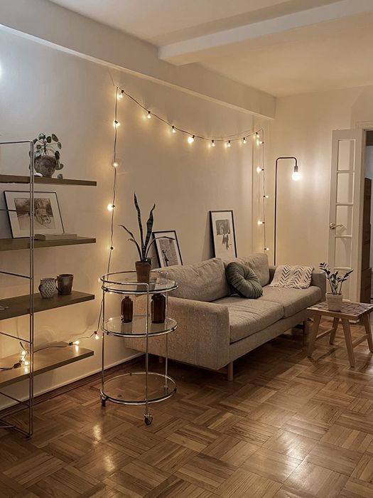 A cozy living room with a grey sofa, string lights, artwork, plants, and a wooden floor. A bookshelf and a small table complement the overall decor.