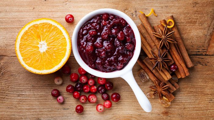 A bowl of cranberry sauce placed on a table alongside an orange half and cinnamon sticks.