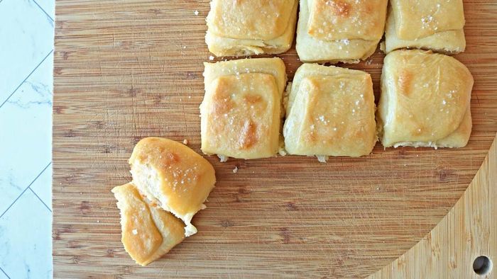 A cutting board holds several dinner rolls, one of which has been torn in half.