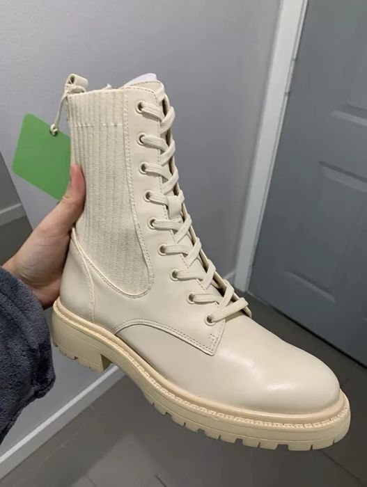 A hand holds a cream-colored, lace-up combat boot, with ribbed fabric around the ankle. The boot is showcased indoors beside a gray door.