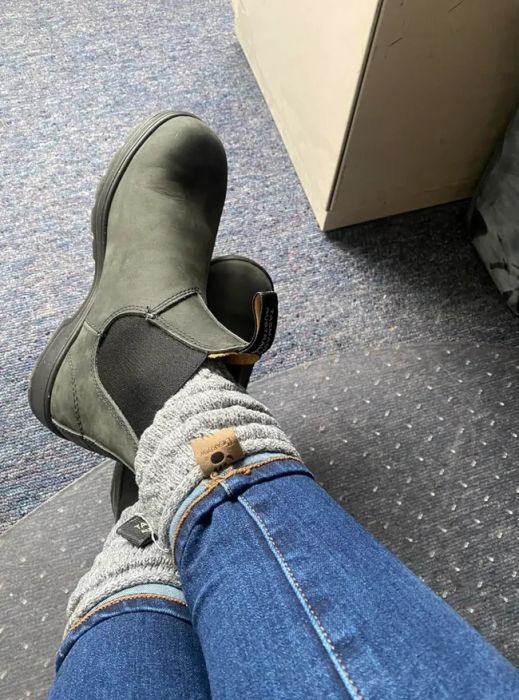 A person wearing gray boots and light gray knitted socks over blue jeans, with their feet resting on a surface. The setting hints at a laid-back shopping moment.