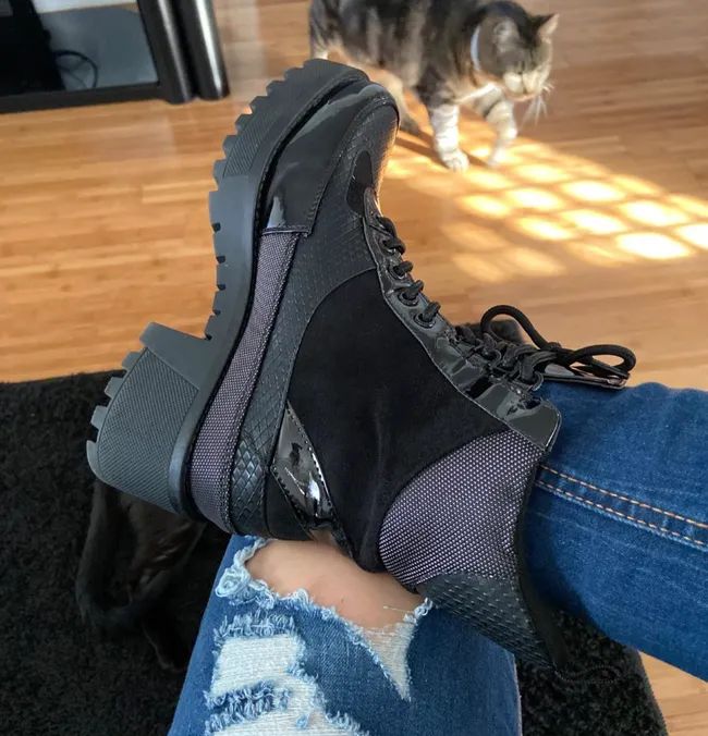 A person is sporting chunky-heeled black boots with laces paired with ripped jeans, while a cat lounges in the background on a wooden floor.