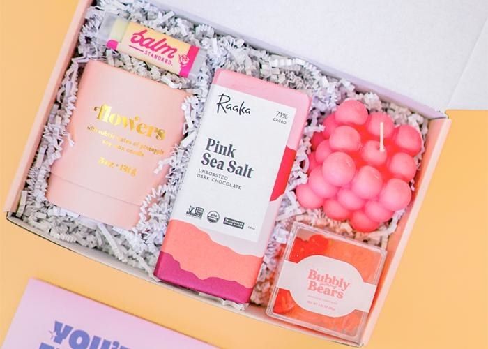 An open gift box containing a lip balm, candle, chocolate bar, another candle, and gummies