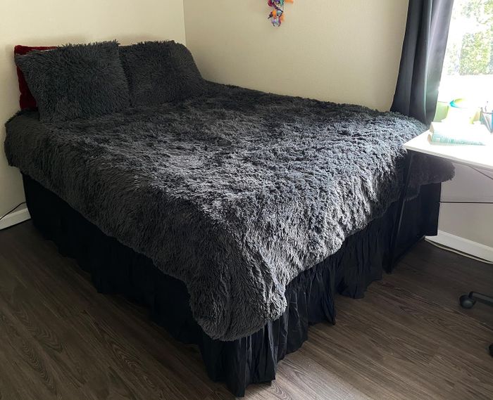 A well-made bed with a soft gray comforter and a black ruffled bed skirt, next to a window and a desk space.