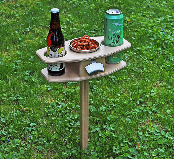 A small table mounted on a stake, featuring two cupholders, a central compartment packed with snacks, and a convenient bottle opener attached.