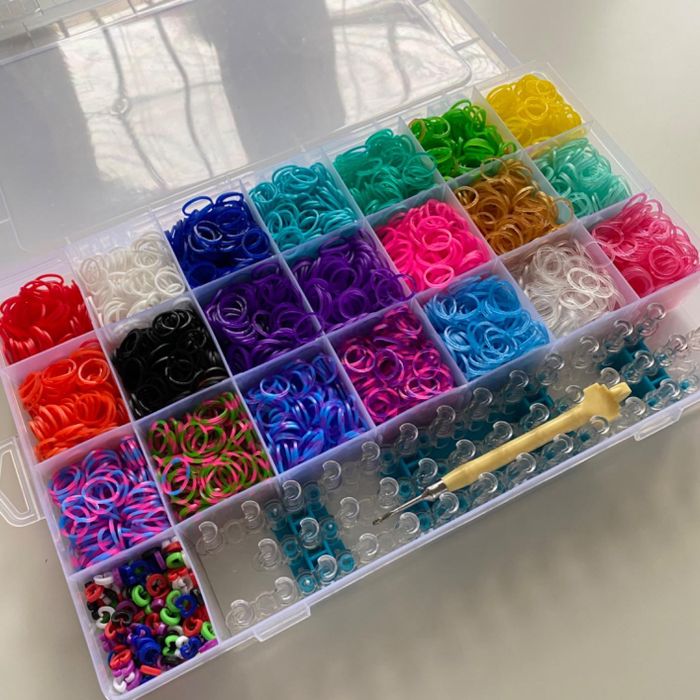 A plastic storage box filled with neatly organized compartments of multicolored loom bands and clear plastic clips, accompanied by a yellow loom tool