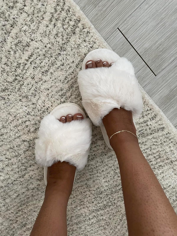 Feet snug in fluffy slippers on a textured rug. A dainty anklet can be seen.