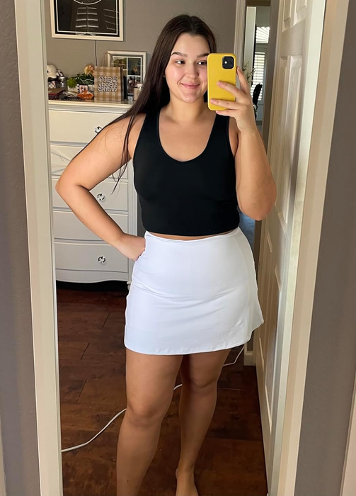 A reviewer taking a mirror selfie, wearing a black tank top and a white tennis skirt.