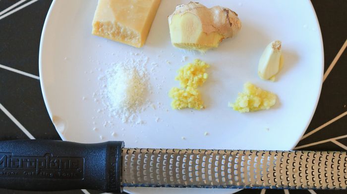 Ginger, parmesan, and a garlic clove on a plate, accompanied by three piles of their grated forms.
