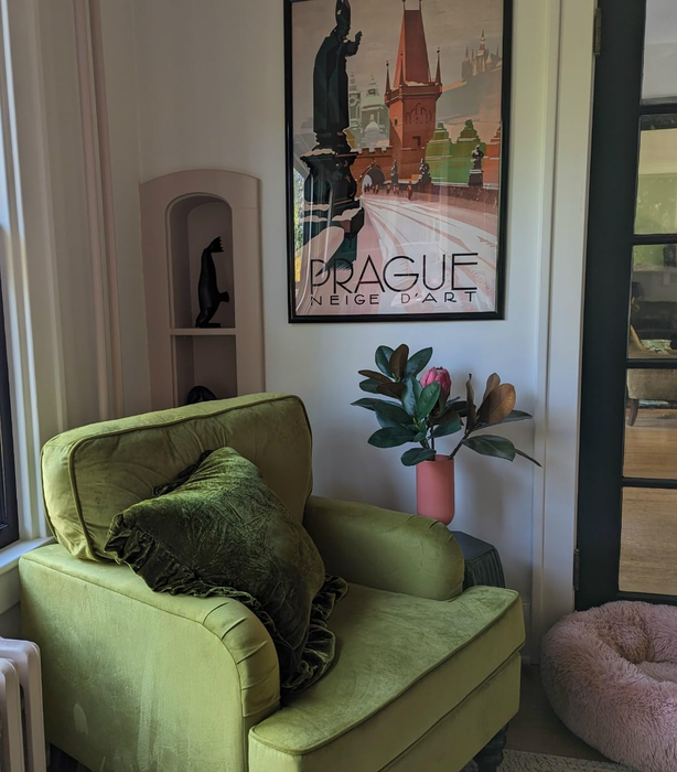 A cozy green velvet armchair with a plush cushion, positioned beside a framed poster of Prague, creating a welcoming atmosphere.