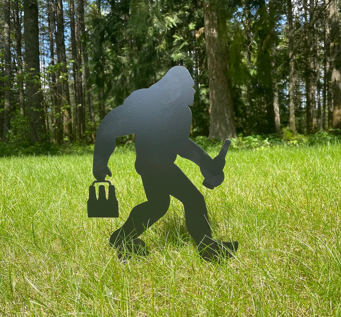 A black metal Big Foot garden stake, outlined and placed in the grass in front of some trees