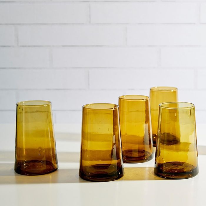 A set of six amber-hued glass tumblers arranged on a white surface, with a brick wall creating a stylish backdrop.