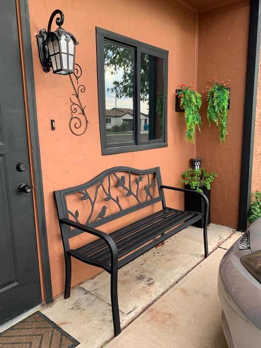A metal bench with an intricate backrest design near the entrance of a house, surrounded by hanging plants